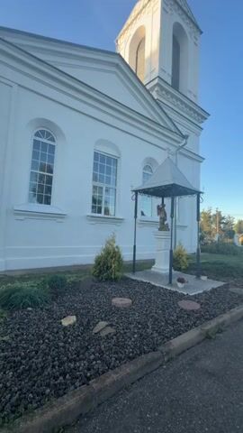 A white church building with a tall bell tower is shown in bright daylight. The camera pans from left to right across the front of the church. In front of the church is a small garden area with a statue of an angel under a small gazebo-like structure. The garden is covered in dark mulch and has a few small plants and rocks. A sidewalk or road runs along the right side of the frame. The sky is clear and blue.