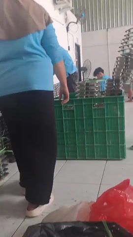 A group of people, mostly women wearing blue shirts and headscarves, are working in what appears to be a food processing or packaging facility. They are handling stacks of metal trays filled with food items. The trays are being placed into large green plastic crates. The environment is industrial, with a tiled floor and metal walls. There are fans and air conditioning units visible. The lighting is bright and artificial.