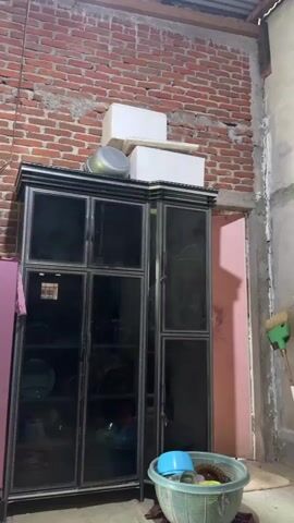 The video shows an indoor scene, likely a kitchen or storage area, with unfinished brick walls and a corrugated metal ceiling. In the center, a tall, dark cabinet with glass doors stands against the brick wall. On top of the cabinet are two white styrofoam boxes and a metal pot. To the left of the cabinet is a pink refrigerator with a floral design. In front of the cabinet is a green plastic basket containing various items, including a blue lid. A colorful rug with circular patterns is visible on the floor in the foreground. The room appears cluttered and utilitarian.