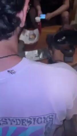 The video shows a close-up view of the back of a person wearing a light purple t-shirt with the words "STUSSY DESIGNS" printed on it. A small, brown grasshopper is perched on the back of the person's t-shirt, near the neckline. The person has dark hair and is wearing a beaded necklace. In the background, there is a wooden table with various items on it, including a bottle of ketchup, a plate with food, and other objects that are partially obscured. Other people are seated around the table, but their faces are not clearly visible.