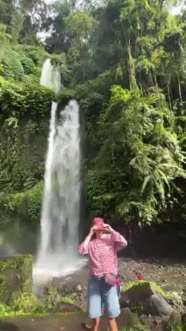 A video shows a woman posing in front of a waterfall. The woman is wearing a red baseball cap, a striped pink and white shirt, and denim shorts. She is standing on a concrete area covered with moss. In the background is a large waterfall surrounded by lush, green vegetation. The waterfall has several distinct streams. Another person enters the frame and takes a photo of the woman with a smartphone. Other people are visible in the background.