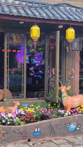 A static shot captures a storefront with traditional Chinese architecture. Two yellow lanterns hang from the eaves above a large window that is open, revealing a musician playing a guitar inside. The interior is dimly lit with colorful purple and blue lights reflecting off the glass. Outside, a flower bed is filled with a variety of colorful flowers. Two deer statues are placed in the flower bed, one larger in the foreground and a smaller one in the background. Below the flower bed is a decorative wall with painted animal figures.