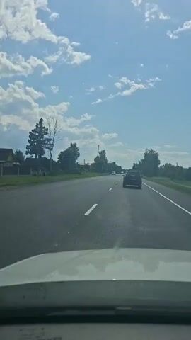 The video captures a first-person view from inside a vehicle driving on a paved road. The road has a white dashed line marking the lane. On the left side of the road, there is a grassy shoulder with trees and houses. On the right side of the road, there are more trees and power lines, and the road continues into the distance with a clear blue sky and scattered white clouds above. Several other vehicles are visible on the road ahead, also traveling in the same direction. The interior of the car, specifically the dashboard, is visible in the foreground. The overall impression is that of a daytime drive through a rural or suburban area.