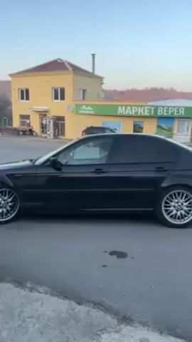 The video shows a black BMW E46 sedan parked on the side of a road. The car has aftermarket wheels. Behind the car is a yellow building with an attached store. The store's sign is in Bulgarian and translates to "Market Vereya Konkosor". The sky is clear and blue.