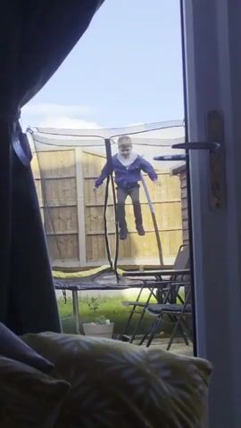 A young person is shown on a trampoline in a backyard, through an open doorway. The subject, wearing a blue top and dark trousers, initially stands on the trampoline. They then lie down and raise their legs. The trampoline is surrounded by a safety net. A wooden fence serves as the backdrop. A table and folding chairs are also visible near the trampoline. Inside, a yellow leaf-patterned pillow is in the foreground. The sky is blue with some clouds.