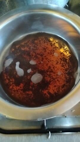 The image shows a top-down view of a metallic pot, likely a saucepan, containing a dark reddish-brown liquid with numerous small, dark particles resembling tea leaves. Several irregular-shaped white foam bubbles are visible on the surface of the liquid. The liquid appears to be hot or recently heated, suggesting it is in the process of being boiled or brewed.

The pot is round and metallic, possibly made of stainless steel or aluminum, with a shiny, reflective inner surface, especially around the rim. Part of a metallic stovetop element or burner is visible to the right of the pot, indicating an indoor kitchen setting.

The overall scene depicts the preparation of a beverage, most likely tea, given the appearance of the liquid and the dark particles. The small, granular nature of the particles is consistent with loose leaf or granular black tea. There are no people visible in the frame, and no discernible text. The provided context indicates this scene takes place in Kikuyu, Kenya.