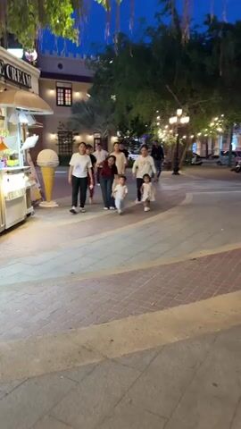 The video shows a group of people walking along a paved walkway in what appears to be an outdoor plaza or street. The lighting suggests it is evening or twilight.

On the left side of the frame, there is an ice cream stand with a large ice cream cone display. A few people are visible near or at the ice cream stand.

A group of approximately seven people, including adults and young children, are walking towards the camera from further down the walkway. They are casually dressed.

The walkway is paved with brick-like patterns. Trees with string lights are present on both sides of the walkway, creating an illuminated pathway. In the background, there are buildings with architectural details, some with illuminated windows. The overall environment appears to be a recreational or commercial area designed for pedestrian traffic.