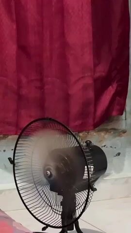 A black oscillating fan is on and blowing air. The fan is situated in front of a red curtain and a textured wall. The fan's blades are blurred from motion.