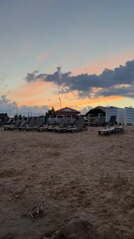 A beach scene at dusk. Rows of empty lounge chairs are set up on a sandy beach. Behind the chairs, there are a few small structures with roofs, and some trees. The sky is filled with clouds, tinged with orange and pink from the setting sun. The overall impression is peaceful and serene.