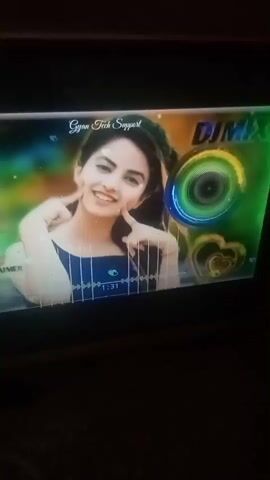 A vertical shot shows a television screen displaying a music playlist. The screen is rectangular and slightly angled. The playlist is set against a colorful background with a woman at the center. She has light skin, dark hair, and is wearing a dark blue top. She's smiling and posing with her fingers pinching her cheeks. Text on the screen reads "Gyan Tech Support" and "DJ MIX." A digital equalizer is visible below the woman, and animated graphics include swirling circles and heart shapes. The room is dimly lit, with the television screen as the main light source. The recording seems to be taken by someone holding a phone close to the TV.
