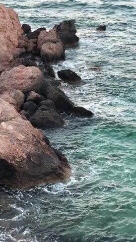This is a high-angle, close-up, outdoor, vertical video of a rocky coastline. The water is a deep turquoise color with white waves crashing against the dark rocks and the larger, reddish-brown rocks on the left side of the frame. The waves are moderately sized and appear to be moving towards the shore. There are no people or animals visible in the video. The lighting suggests it is daytime, possibly overcast.