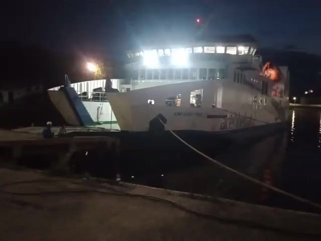 The video shows a ferry docked at night. The ferry is white and well-lit. Its name, "KMP CAKRA NUSA", is visible on the side. A rope connects the ferry to the dock. The water is dark and reflects the lights from buildings on the opposite side. There are some clouds in the sky, illuminated by the twilight. On the right side of the frame, people can be seen walking on the dock. Some greenery is visible along the edge of the dock where the camera is located.