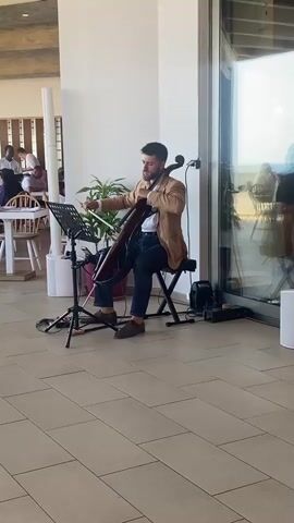 A male cellist plays in a modern indoor space with large windows. He is seated on a black stool, wearing a tan jacket, white shirt, and dark pants. His cello is dark red, and he holds the bow with his right hand, playing with concentration. A music stand is positioned in front of him, holding sheet music. Behind the cellist, there are people seated at tables, suggesting a restaurant or event space. The floor is tiled with a light, sandy color. To the right of the cellist, there's a large window with a view of the outdoors, and reflections of people and the environment are visible on the glass. The space appears bright and airy.