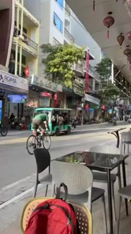 The camera is positioned under the awning of a cafe in Vietnam, showcasing a street bustling with activity. Tables and chairs are visible, with a red backpack resting on one of the chairs.  A cyclist rides down the street, passing a small green electric bus carrying passengers.  Motorbikes weave through traffic. The street is lined with buildings, adorned with signage in Vietnamese and colorful lanterns.  People can be seen walking on the sidewalks and sitting at other cafes across the street.  The scene depicts a typical day in a vibrant Vietnamese city.