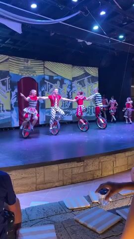 The video shows a performance on a stage. Four people in colorful, patterned costumes are riding unicycles. Two individuals in red and white striped shirts and black pants are on the left and right. In the center, two individuals are wearing red tops and patterned skirts. The backdrop of the stage features a large, yellow mural with a central red curtain. The performers are holding hands and moving in a synchronized manner across the stage. The lighting on the stage is blue and purple. In the foreground, the audience is visible, with some people sitting and others standing, with what appear to be cushions or mats on the floor in front of them.