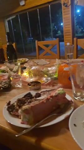 The video shows a close-up of a person's face, with their eyes wide and looking directly into the camera. The person is wearing a white, collared shirt. The background is blurred, but appears to be an indoor setting with dim lighting. There are also brief glimpses of a table laden with leftover food and dishes, suggesting a meal has recently concluded. Some of the food visible includes cake, what appears to be a pumpkin, and various other items on plates. The overall impression is of a candid, perhaps slightly surprised or playful moment captured at the end of a gathering.