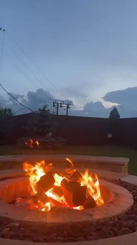 A wide shot captures a backyard scene as dusk settles. In the foreground, a circular fire pit radiates intense orange flames and glowing embers. The pit is constructed of stone and surrounded by decorative dark stones. Beyond the fire pit, a manicured green lawn stretches back to a dark wooden fence. Several pine trees and other greenery are visible behind the fence, with utility poles and wires silhouetted against a cloudy, twilight sky. The overall impression is of a serene evening outdoors.
