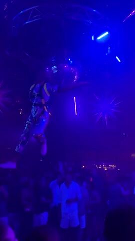 A nighttime aerial performance takes place in a dimly lit nightclub. A female performer in a white leotard is suspended in the air by two white silk fabrics. She is positioned in the center of the frame, her body gracefully contorted as she performs aerial maneuvers. The nightclub is filled with a crowd of people, many of whom are dressed in white. The environment is illuminated by vibrant purple and green lights, with disco balls and star-shaped light fixtures hanging from the ceiling. The overall atmosphere is one of excitement and celebration, with a focus on the aerial artistry.