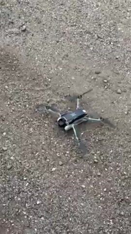 A small drone lifts off from a gravel surface. The drone is black and white, with four propellers. The ground is composed of gray gravel. The drone rises steadily.
