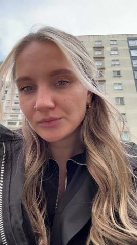 A woman with long, blonde hair, a nose ring, and wearing a black jacket looks directly at the camera. Her hair is slightly windblown. The background features a tall apartment building with multiple rows of windows under a cloudy sky. The camera angle is low, looking slightly up at the woman.