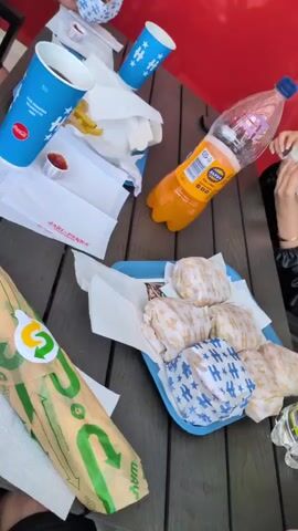 A static, top-down view shows a dark wooden table laden with various fast-food items. On the right, a blue plastic tray contains five individually wrapped food items, resembling burgers or sandwiches, with some wrappers being plain white and others blue with white stars. Adjacent to the tray, a person's hand, wearing a dark sleeve and a bracelet, is visible near a wrapped item. To the left, a Subway paper bag lies horizontally. Further left, two white paper bags are present; one contains visible french fries and a small foil cup of red sauce. Two blue cups, one larger with a Coca-Cola logo and one smaller with a star pattern, are positioned near the paper bags. A large clear plastic bottle filled with orange liquid stands on the right side of the table. A red wall is visible in the background.