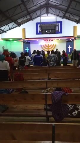 Inside a church, a group of about 20 people are dancing in front of the altar. The altar has a large menorah in the center, flanked by crosses on each side. Above the menorah is a screen under a sign that says "Valley Church." The church has a metal roof and rows of wooden pews that are mostly empty. Some clothing items are on the pews. There are some colored lights pointing at the altar. The overall feel is vibrant and energetic.