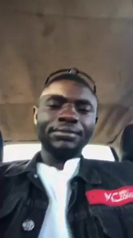 The video shows a man sitting in a car. He's wearing a black jacket over a white shirt. He's looking directly at the camera and speaking. He puts on sunglasses. The interior of the car is visible in the background, including the ceiling and headrest.