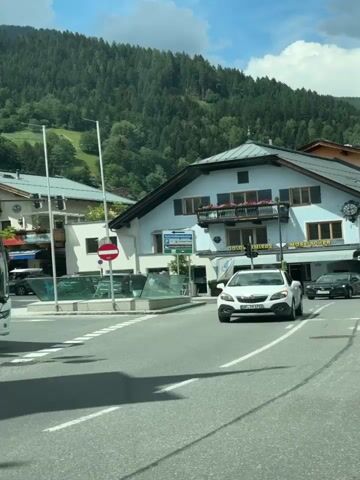 A white SUV with a bicycle rack on the back drives forward in the foreground. A black sedan follows behind it. A light blue building with a balcony is visible in the background, along with a mountain covered in trees. A street sign is also visible.