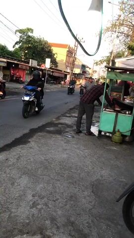 The video shows a street scene in what appears to be a Southeast Asian country, possibly Indonesia, based on the architecture and the presence of motorbikes. The time of day is late afternoon or early evening, as indicated by the warm lighting and long shadows. The street is paved with asphalt and shows signs of wear, with some water stains and debris.

In the foreground, on the right side of the street, there is a food cart. A man in a plaid shirt and dark pants is leaning over the cart, possibly preparing food or serving customers. Next to the cart, a green propane tank is visible.

Further down the street, several motorbikes are in motion. One motorbike on the left side of the road is carrying two people, one of whom is wearing a pink hijab. Another motorbike is further ahead, also with a rider. On the right side of the road, a man is standing near another food cart, partially obscured by the foreground cart.

On the left side of the street, there are several small shopfronts with awnings and signs. Trees and utility poles with electrical wires are also present. The sky is visible in the background, with clear weather.

The camera appears to be mounted on a motorbike, as evidenced by the reflection of a mirror in the upper right corner of the frame and the slightly shaky perspective. The overall atmosphere is one of everyday street life, with commerce and transportation taking place.