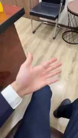 Here's a description of the video:
The video shows a person sitting down, wearing a navy suit and a white-collared shirt. Their left hand is extended, palm facing up, in front of their lap. The setting appears to be an office, with a wooden desk to the left, a black office chair, and a small side table with a wire base. The floor is light-colored wood laminate. The person's right leg, clad in navy pants and a black shoe, is also visible.