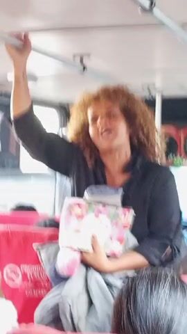 The video shows a woman standing in a moving bus, holding onto a safety bar with her left hand and clutching an assortment of items, including a pillow, blanket, and a floral-patterned case, in her right arm. Her hair is curly and brown, and she wears a dark long-sleeved shirt. The bus interior has red seats, with other passengers visible seated around her. There are signs in Spanish on the bus as well.