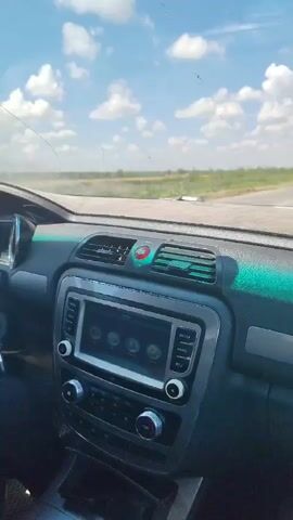 The video is shot from inside a car looking out through the windshield at a road and sky. The car's dashboard is visible in the foreground, with a touchscreen radio and air vents. A white truck is seen briefly driving past the car on the road. The sky is blue with scattered white clouds, and the landscape outside appears to be rural or undeveloped.