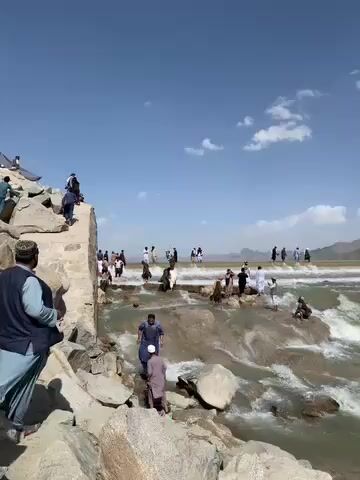 The video captures a wide shot of a scenic outdoor location, likely a river or a natural waterfall with numerous people present. The scene is dominated by a flowing body of water that cascades over natural rock formations and a man-made structure, creating small rapids and foamy white water.

In the foreground, to the left, a large rocky bank is visible, with several people standing or sitting on the rocks. One man in a light blue traditional outfit (shalwar kameez) and a dark vest stands facing away from the camera, looking towards the water. Another man, also in traditional attire, is visible slightly behind him on the rocks.

The midground is bustling with activity. Dozens of people, predominantly men and boys, are scattered across the rocky expanse of the water feature. Some are wading in the shallow parts, others are sitting on the rocks close to the water, and many are simply standing and observing. Most individuals are dressed in traditional clothing, often light-colored shalwar kameez. A visible stone structure, possibly part of a weir or dam, stretches across the water, and people are walking on or alongside it. The water appears swift-moving and turbulent in places, especially where it cascades over drops.

In the background, the water extends further, appearing calmer in the distance before meeting a distant, hazy landscape of hills or mountains. The sky is bright blue with a few scattered white clouds, indicating clear and sunny weather. The overall impression is that of a popular recreational spot where people are enjoying the natural environment and the water.

The video also includes ambient audio of rushing water, enhancing the immersive feel of the scene. No text is visible in the video.