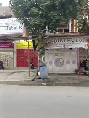 A video shows a street scene with several businesses and people on motorcycles. In the first frame, a building with two storefronts is visible. One storefront is labeled "Bee Cosmetics" and the other is labeled "APOTEK Jabal Rezki." A person on a motorcycle is in front of the "Bee Cosmetics" store. The second frame shows a small building with a red gate and a kiosk with a white and silver exterior. The third frame shows more storefronts, with one having pink signage and the other appearing to be a food stall. The fourth frame shows a blue van with "BSI" written on it, possibly a mobile banking unit, with a person standing near it and two motorcycles parked nearby. The fifth frame shows a building with a red roof and trees in front of it. There are two tents set up on the sidewalk, one green and one yellow, with tables and chairs under them. A motorcycle is parked near the yellow tent. The video is shot from a moving vehicle, likely a car or another motorcycle, and the motion causes some blurriness. The overall environment appears to be an urban or suburban street with commercial establishments. The sky is overcast.
