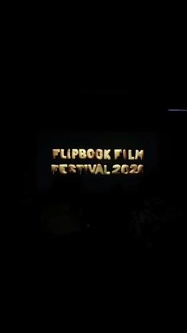 This video captures a brief moment in a darkened room, likely a cinema or screening hall. The primary focus is a projection screen displaying the words "FLIPBOOK FILM FESTIVAL 2020" in a stylized, three-dimensional font with a textured, golden appearance. The text is illuminated against a dark, possibly blue, background. In the foreground, silhouetted figures of people are visible from behind, seated and facing the screen. Their presence suggests an audience watching the presentation. The overall atmosphere is one of anticipation or quiet observation, characteristic of a film screening event.