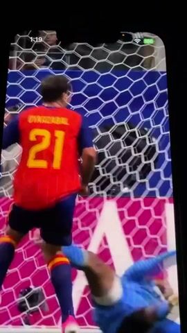 A soccer player in a red and yellow uniform, with the number 19 on his back, smiles as he jogs in front of a soccer goal with a blue and pink net. A goalkeeper in a blue uniform lies on the ground near the goalpost. The scene is captured from a phone screen, with a timestamp of 1:19 visible in the top left corner. The background shows a stadium with a large crowd and advertisements. The video then cuts to a different angle, showing the same player running on a green soccer field, celebrating. The rest of the video depicts a soccer game in progress, with players in red and white uniforms actively playing around the goal. One player in a red and yellow uniform is seen kicking the ball, while another player in white tumbles to the ground.