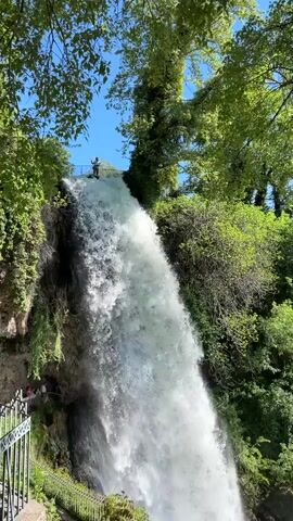 The video shows a wide, tall waterfall, lush green foliage, and a bright blue sky. A metal railing runs along a pathway at the bottom of the frame, with a few people visible beyond it, but too far to identify. On top of the waterfall, a person stands at the edge of a platform, appearing to take in the view. The water rushes down the rocky cliff, with mist rising at the base. The dense vegetation surrounds the waterfall, creating a vibrant green backdrop. The scene conveys a sense of natural beauty and wonder.