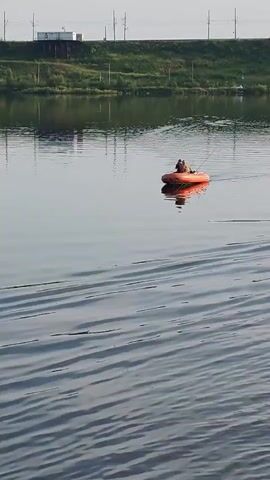 The video shows a wide shot of a body of water, likely a river or lake. The water is calm with gentle ripples, reflecting the sky and the distant shore. On the far bank, there is a strip of green vegetation, possibly trees and bushes, with a railway line running along it. A white structure, possibly a building or train car, is visible on the railway line. In the foreground, the water is darker and more textured due to close-up ripples. In the middle of the frame, an orange inflatable raft is moving across the water from left to right. Two people are visible in the raft, both appearing to be male. They are sitting and facing away from the camera. One person is holding what appears to be a fishing rod, suggesting they are fishing. The raft is creating a small wake behind it. The sky above is clear and pale, indicating it is likely daytime, possibly early morning or late afternoon given the soft light. The overall atmosphere is peaceful and serene.