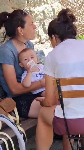 A close-up shot shows three people on a wooden bench outdoors. On the left, a woman with dark hair pulled back in a bun, wearing sunglasses on her head and a dark grey t-shirt, holds a baby in her lap. The baby, wearing a white t-shirt with yellow writing, is drinking from a clear bottle with a white nipple. The baby's face is turned towards the camera. To the right of the woman and baby, another person, viewed from the back, is wearing a white t-shirt and dark burgundy shorts. They are seated on a wooden chair with a metal frame and wooden slats. The background consists of a stone wall and a netted fence. The overall lighting suggests a sunny day.