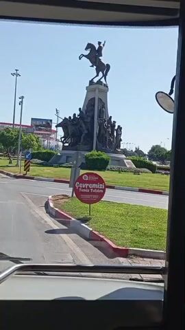 The video shows a view from inside a vehicle, looking out at a large equestrian statue. The statue depicts a figure on horseback with a group of other figures surrounding the base. The monument is situated on a grassy roundabout, with a red and white striped curb. A circular red sign with white text is placed in the grass in front of the monument. The background shows trees, streetlights, and buildings, suggesting an urban environment. The sky is clear and blue.