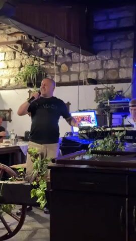 A bald man wearing a black t-shirt with a logo and tan shorts is singing into a microphone at an indoor event. He is standing in front of a table with a cart filled with plants, and a DJ setup with keyboards and a laptop is visible behind him. The room has stone walls, wooden beams, and hanging plants. There are other people seated at tables in the background. The lighting shifts from white to purple.
