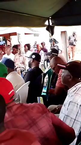 A group of people are seated under a tent. Some people are wearing hats. There is a wall in the background.