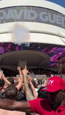 The video shows a large crowd with their hands in the air at an outdoor party or concert. There is a stage in the background with a large digital display above it.  Smoke or fog effects are being used, and white confetti is falling. A man in a red LIV Beach hat and a red shirt is facing the camera in the foreground. The stage has the name “DAVID GUETTA” prominently displayed.