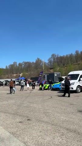 The video shows a crowd of people gathered in a large, paved parking lot under a clear blue sky. Several cars, including a bright green sports car and a blue sports car, are parked in the lot, along with other vehicles. There are tents and banners set up in the background, and a line of shipping containers is visible along the edge of the lot. A treeline is behind the parking lot. People of all ages are walking around, some in groups, while others are alone. A small child walks with an adult. The overall impression is that of a public event or gathering, possibly a car show.