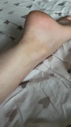 A close-up video shows a person's foot and lower leg on a patterned bedsheet. The foot is positioned at an angle, with the heel visible on the left side of the frame and the toes extending towards the right. The skin appears pale, with some subtle shading and texture. The bedsheet has a repeating pattern of light brown hearts. The lighting suggests an indoor environment, possibly a bedroom.