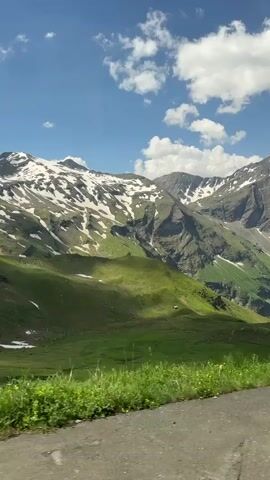 The video shows a panoramic view of a mountainous landscape under a blue sky with scattered white clouds. The foreground features a grassy verge with wildflowers and a rough surface that could be a roadside or trail. A wooden post stands prominently on the right side of the frame. Beyond the foreground, rolling green hills descend into a valley. Further in the distance, large, rugged mountains rise, their peaks capped with snow. Sunlight casts shadows across the landscape, highlighting the contours of the hills and mountains. There are no people or visible actions taking place. The environment is natural and appears to be an alpine or mountainous region.