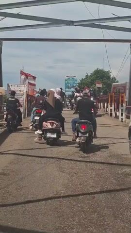 A group of people on motorcycles are riding across a bridge under a cloudy sky. Indonesian flags are visible on the sides of the bridge. Some of the motorcyclists are wearing helmets, while others are not. A sign with the number 30 is visible on the right side of the bridge. The bridge appears to be made of metal and concrete. The road surface is asphalt.