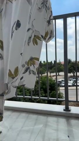 The video shows a close-up of a white garment with a floral pattern hanging in the wind on a balcony. The garment is white with a pattern of green, pink, and gray flowers and leaves. The garment is blowing in the breeze. In the background, there is a metal railing, a tiled floor, a street with parked cars, trees, and a blue sky with white clouds.