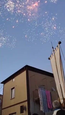 The video shows a clear blue sky with fireworks exploding. In the foreground, there is a tan building with a dark brown roof and trim. There is a balcony on the building with laundry hanging on a clothesline. A white banner is attached to a pole on top of the building. In the background, another building with arched windows is visible. The fireworks are colorful and appear to be part of a celebration.