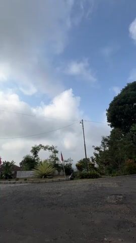 The video begins with a view of a cloudy sky and a dirt road. As the camera moves forward, an arched entrance gate comes into view. The gate is painted yellow and green and is decorated with red and white flags. There is a small yellow booth to the left of the gate, possibly a guardhouse. A motorcycle is parked in front of the booth. In the background, there are trees and buildings. The camera continues to move forward, showing the entrance to a property. The flags are clearly visible, as are several motorcycles parked on the side of the road. The video ends as the camera passes through the gate.